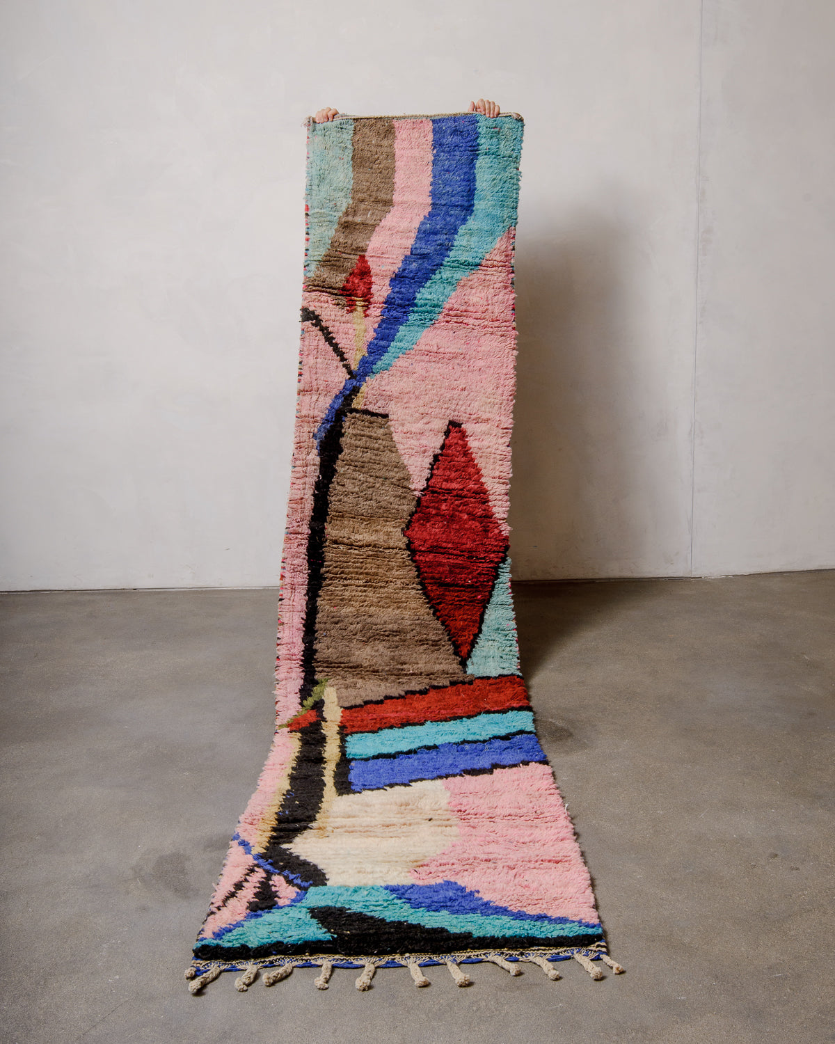 Berberlin vintage Berber runner rug made of colourful wool, being held up in front of a white wall and concrete floor.
